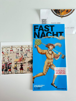 Flyer mit der Aufschrift 'FASTNACHT' und einer Illustration einer historischen Figur in bunter Kleidung mit Federhut, daneben eine Postkarte mit einer historischen Tanzszene und ein Museumseintrittsticket