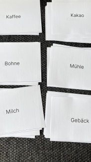 Mehrere weiße Karten mit den Wörtern 'Kaffee', 'Kakao', 'Bohne', 'Mühle', 'Milch' und 'Gebäck' liegen auf einem dunklen, strukturierten Untergrund.