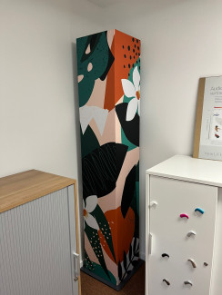 Den zentralen Bereich des Bildes dominiert eine hohe, zylindrische Säule oder einen Lautsprecher, der mit einer Stoffhülle mit einem abstrakten Muster bedeckt ist. Das Muster besteht aus stilisierten Blumenmotiven und geometrischen Formen in einer harmonischen Farbpalette aus Orange, Grün, Weiß und Schwarz, die einen visuellen Kontrast zum neutralen Hintergrund bilden. Am linken und rechten Bildrand ist jeweils eine Aufbewahrungslösung platziert.