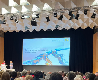 Blick von hinten auf einen voll besetzten Vortragssaal mit vielen sitzenden Personen. Im Vordergrund ist eine große Leinwand zu sehen, auf der eine Präsentationsfolie mit dem Titel 'Demenz in unterschiedlichen Lebensphasen' angezeigt wird. Zwei Personen stehen auf der Bühne links neben der Leinwand. Die Decke ist mit geometrischen, weißen Elementen gestaltet. Die Wände sind holzvertäfelt.