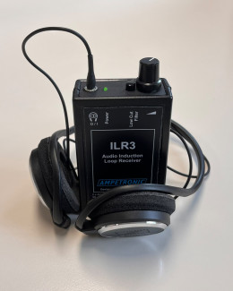 Ein rechteckiges, schwarzes Gerät mit der Aufschrift 'ILR3 Audio Induction Loop Receiver' steht auf einer hellen Oberfläche. Oben am Gerät befinden sich ein Drehregler, ein kleiner Schalter und eine grüne Kontrollleuchte. Ein schwarzes Kabel ist eingesteckt. Um das Gerät liegt ein Paar ohrumschließender Kopfhörer mit gepolsterten Ohrmuscheln und silberfarbenen Außenseiten. Das Markenlogo 'Ampetronic' ist auf der Vorderseite des Geräts sichtbar.