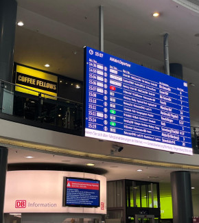 Große digitale Abfahrtstafel mit Zugverbindungen und Uhrzeit über einem DB-Informationsschalter in einem Bahnhof.