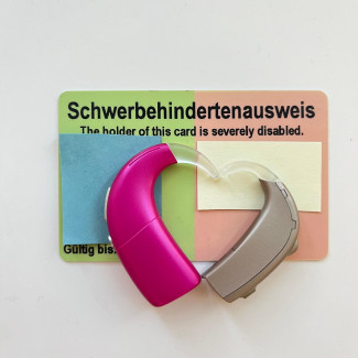 Ein Schwerbehindertenausweis mit der Aufschrift Schwerbehindertenausweis. The holder of this card is Beverly disabled. liegt auf einer hellen Oberfläche. Auf dem Ausweis sind bestimmte Details durch farbiges Papier unkenntlich gemacht. Auf dem Ausweis liegt ein Pinkes Cochlea-Implantat-Implantat sowie ein silbernes Hörgerät. Beide sind so zusammengelegt, dass sie mit ihren Winkeln eine Herzform bilden.