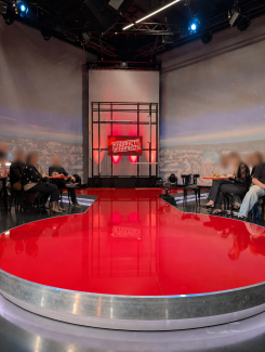 Ein Fernsehstudio mit einer Kabarettbühne: Im Vordergrund befindet sich eine große, runde, glänzend rote Plattform. Links und rechts sitzen mehrere Zuschauerinnen und Zuschauer an kleinen Tischen. Im Hintergrund ist eine erhöhte Bühnenkonstruktion mit einem Bildschirm zu sehen, auf dem „Kabarett Franken“ steht. Die Bühne ist in Rotlicht getaucht, darüber hängen Studio- und Scheinwerfertechnik an einem Gestell. Im Hintergrund ist eine stilisierte Stadtkulisse als Wandbild erkennbar.