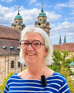 Porträt einer lächelnden Frau mit kurzen weißen Haaren und Brille. Sie trägt ein blau-weiß gestreiftes Oberteil mit einem angesteckten Mikrofon. Im Hintergrund sind die Türme der Nürnberger Egidienkirche vor einem leicht bewölkten, blauen Himmel zu sehen