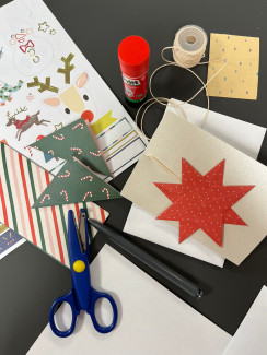 Bastelmaterialien auf dunklem Untergrund: Schere, Klebestift, Faden, Papier mit Weihnachtsmotiven, roter Stern aus Papier mit weißen Punkten, weiße und schwarze Blätter.