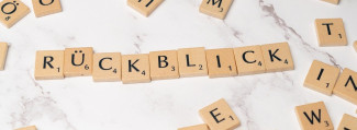 Das Bild zeigt Scrabble-Spielsteine, die das Wort "RÜCKBLICK" bilden. Die Buchstabensteine liegen nebeneinander auf einer weißen Oberfläche mit leichter Marmorierung. Einige weitere Scrabble-Steine liegen lose um das Hauptwort herum verteilt.